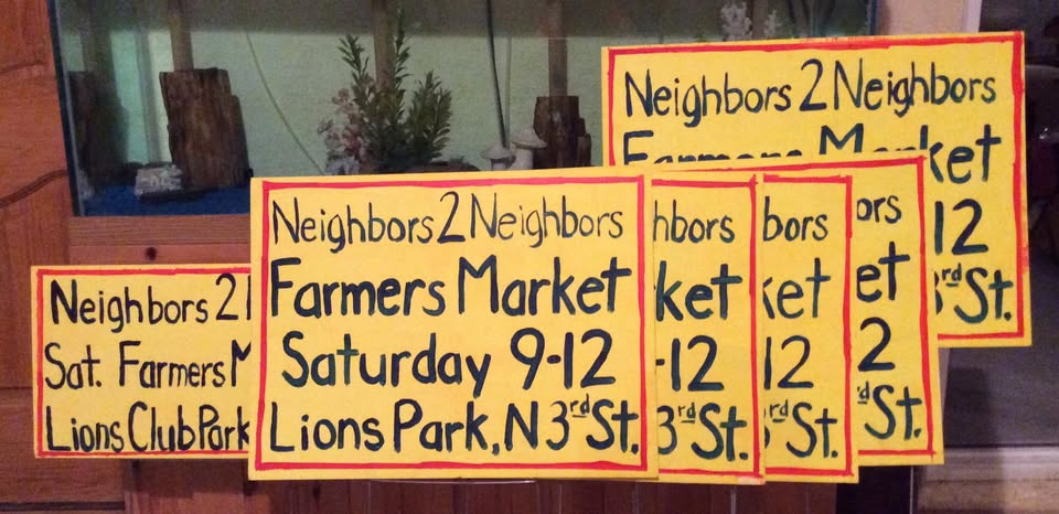 Hand-lettered yellow Neighbors 2 Neighbors Farmers Market signs: Saturday 9–12, Lions Park, N 3rd St.